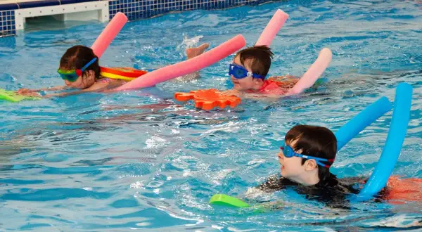 Children in goggles and swim aids enjoy a swim lesson, practicing their strokes in a bright blue pool.