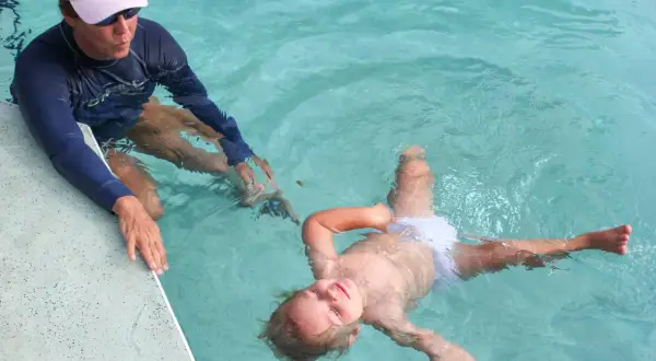 A child floats on their back in a pool, learning to swim. An instructor in a blue rash guard and pink hat watches.