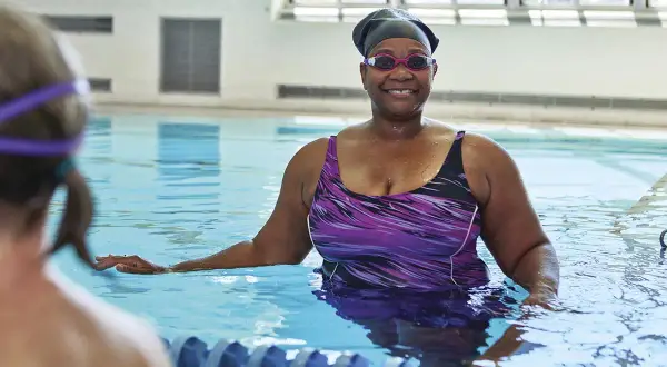 Smiling woman in a purple swimsuit and swim cap enjoys swim lessons for seniors.
