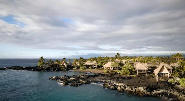 Tropical bungalows on a rocky coast in Kona, Hawaii. The calm ocean reflects the cloudy sky, a stark contrast to the power of a tsunami.