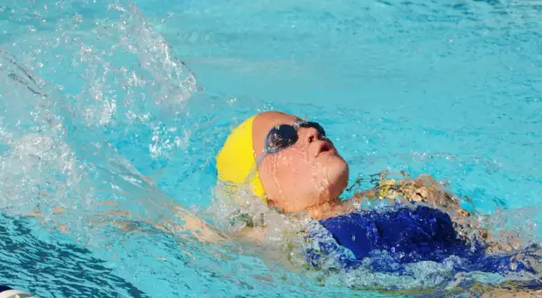 A young swimmer with a yellow cap and goggles performs the back stroke, creating a splash as they move through the blue water.
