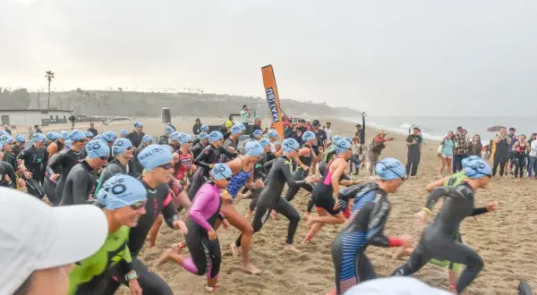 Athletes in wetsuits and blue caps sprint on the sand at the Zuma Beach Triathlon, starting their race with determination.
