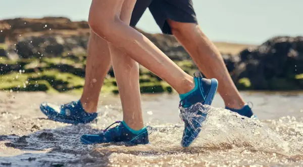 Two people wearing aqua jog shoes splash through shallow water on a rocky beach.