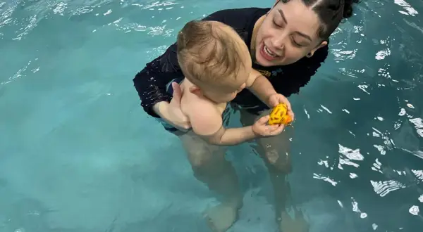 A woman holds a baby in a pool, both smiling. The baby plays with a yellow toy. This scene shows that you can swim with tubes in your ears.