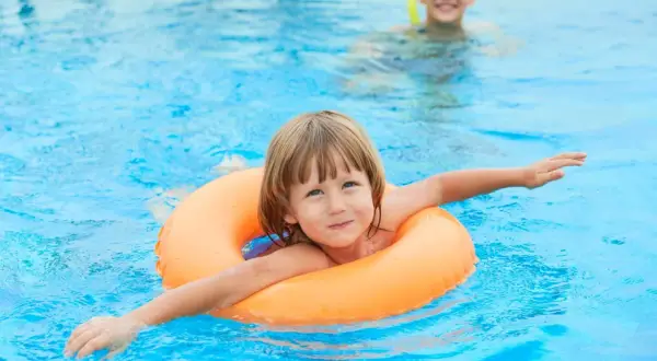 A happy child floats in an orange ring in a pool. Another child in goggles is in the background. This image reminds us when to stop swim lessons: when they're confident and enjoying the water!