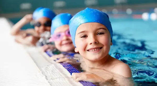 Happy kids in blue swim caps at swim lessons for kids, smiling by the pool edge.