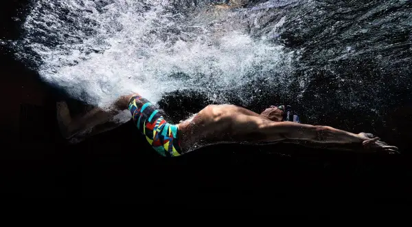 Underwater shot of a swimmer in a colorful tech suit, arms extended, with bubbles and light above.