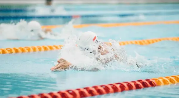 A swimmer in a white cap and goggles powers through the water, creating a splash. Their focused swimmer posture is evident as they propel forward in the lane.