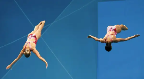 Two divers perform synchronized dives, showcasing their mastery of olympic diving terms as they execute a complex maneuver against a vibrant blue background.