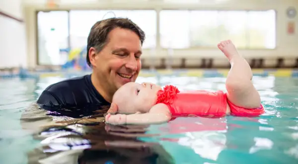A father smiles as he holds his baby in a pool, showing that kids can learn to swim from a very young age.
