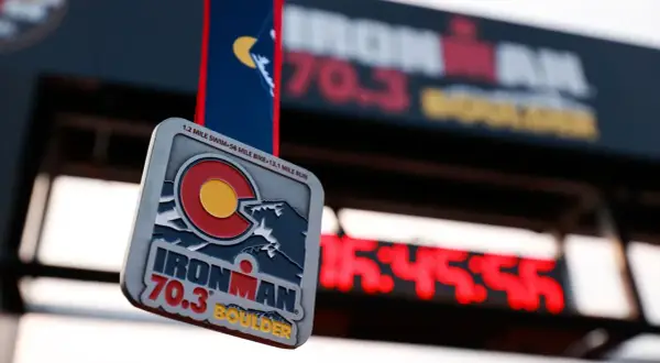 Ironman 70.3 Boulder medal hangs in front of a finish line clock. The medal features the Colorado flag and mountains.