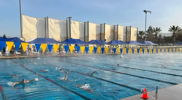 Swimmers race in lanes during a circle swim event. Blue and yellow banners line the pool, with tents and spectators in the background.
