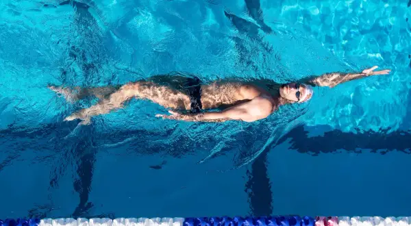 A male backstroke swimmer glides through the bright blue water of a pool, his body streamlined and arms extended.