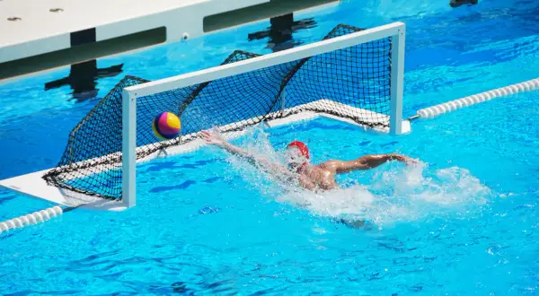 A water polo player in a red cap lunges for the ball near the net, splashing water. Intense underwater games are a thrilling spectacle.