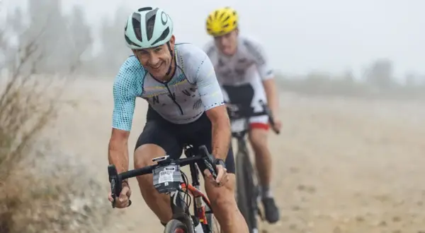 Smiles and determination on the faces of cyclists during a gravel triathlon. The lead rider, wearing a light blue and white jersey, grins as he navigates the dusty course.