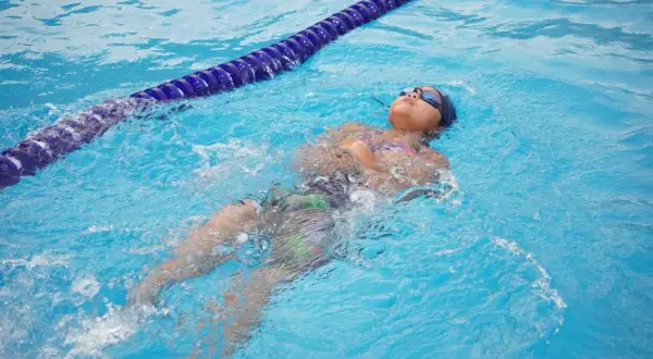A child practices how to tread water in a pool, floating on their back with goggles on.