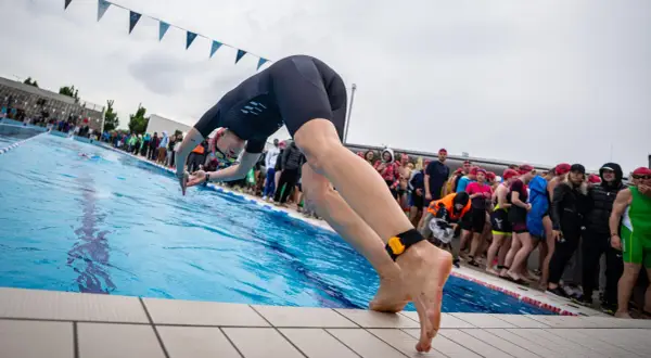 A swimmer in a black wetsuit dives into a pool, ready for advanced swim workouts. Spectators watch from the side.