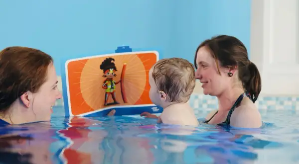A toddler enjoys a fun toddler swim school lesson, looking at a colorful book with two smiling instructors in the pool.
