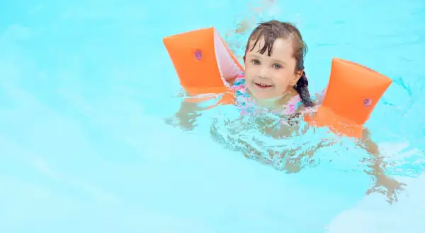 A happy young girl with bright blue eyes is floating in the pool, wearing orange arm floaties and a colorful swimsuit.
