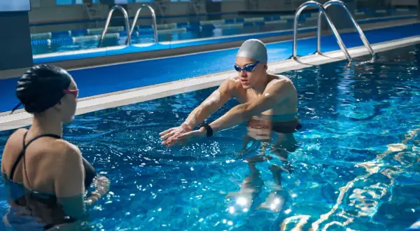 An adult swim coach instructs a swimmer in a pool, demonstrating proper arm stroke technique.