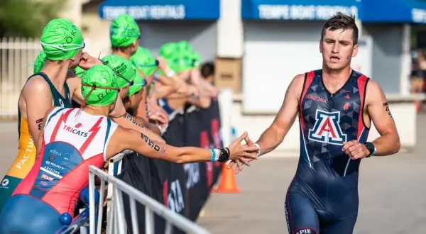 A male NCAA triathlon athlete in a dark blue and red suit with an "A" logo high-fives a competitor.