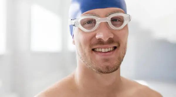 A smiling man in a blue swim cap and goggles, ready to swim. He's wondering, "When can I swim after wisdom teeth removal?