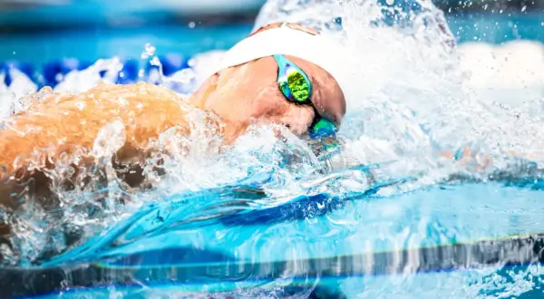 A swimmer's head emerges from the water, goggles reflecting the blue pool. Water splashes around them, a dynamic scene of fun swim drills.