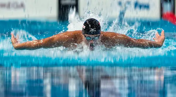 A swimmer in a black cap and goggles performs the butterfly stroke, arms extended, creating a splash as they take a breath.