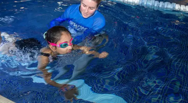 A swim instructor guides a young girl learning to swim. The question of how long does it take to learn how to swim varies, but with practice, she'll be swimming confidently.