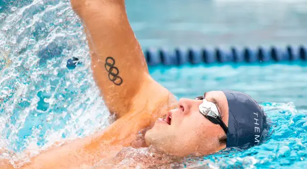 Swimmer in backstroke position, with Olympic rings tattoo on arm, mid-stroke, water splashing.