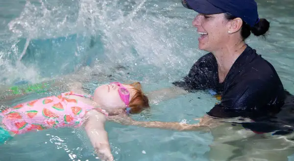 A child in a watermelon swimsuit and pink goggles floats on their back while a smiling instructor helps them practice treading water.