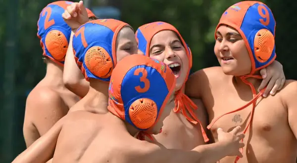 A group of young water polo players, wearing orange and blue caps with numbers, celebrate a victory. One boy, with a big smile, shouts "Habawaba!
