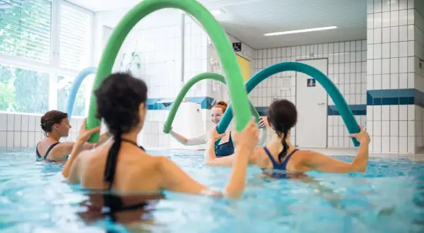 Women enjoy an aqua zumba class, holding colorful pool noodles for resistance and fun in the water.