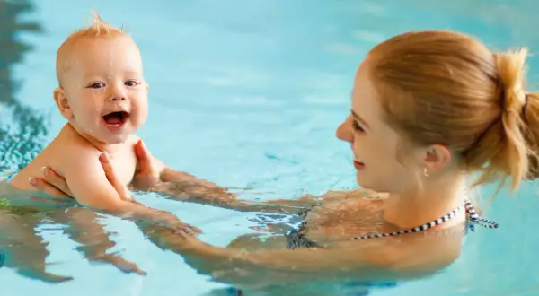 A happy baby laughs during baby and me swim classes, held by their mother in a bright blue pool.