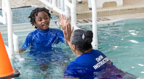 A young boy with dreadlocks in a blue rash guard gives a high-five to a swim instructor. This is a moment of connection during an autism swim lesson.