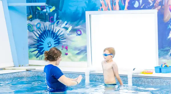 A swim instructor guides a young boy in goggles during 6 month swim lessons. The pool has a fun underwater mural.