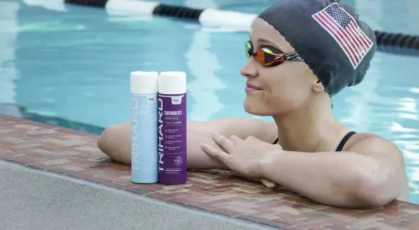 Swimmer with an American flag cap and goggles rests by the pool, with TRIHARD swimmer's shampoo bottles nearby, offering a solution on how to protect skin from chlorine.