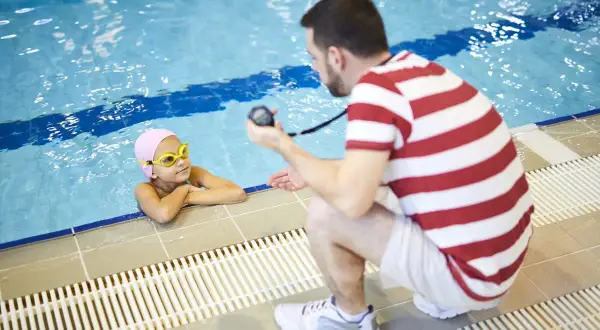 A mobile swim instructor crouches by the pool, holding a stopwatch, as a young girl in a pink cap and goggles rests her arms on the edge.
