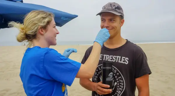 A woman in a blue shirt and gloves gently swabs a man's nose on a beach. He's wearing a cap and a t-shirt that reads "Trestles." This is part of a study to see if you can swim with a staph infection.
