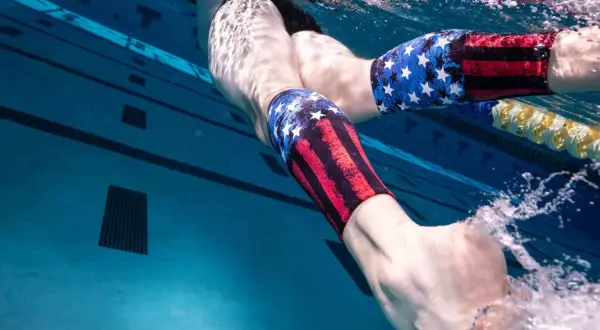 Underwater view of a swimmer's legs in a pool, wearing patriotic-themed compression sleeves. Wondering why do I get foot cramps in the pool?