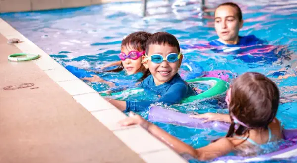Children enjoy swim lesson games, splashing and laughing in the pool with goggles and flotation devices.