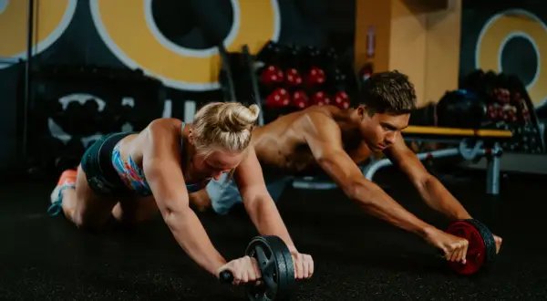 Two athletes, like swimmers training, perform ab roller exercises in a gym.