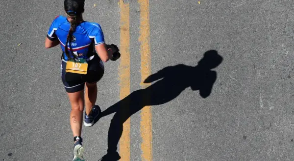A runner in blue triathlon apparel, bib number 107, runs on a road, casting a long shadow.