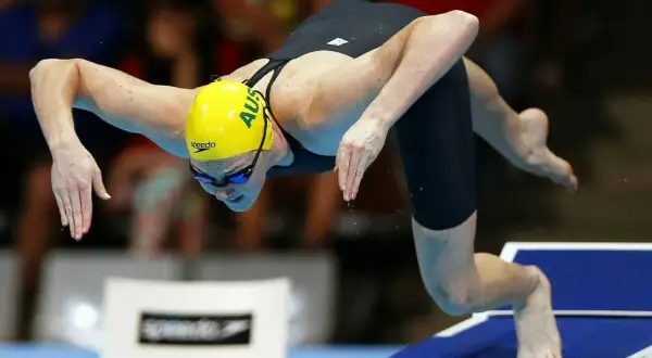 An Australian swimmer in a yellow cap prepares for a powerful high diving start, poised on the edge of the block.