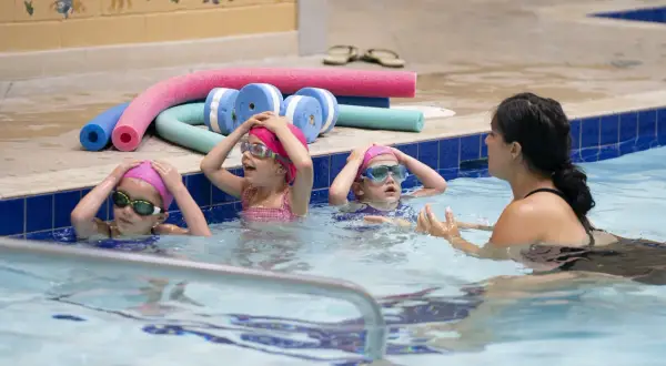 Children in a pool during youth swim lessons, wearing pink caps and goggles, with a swim instructor.
