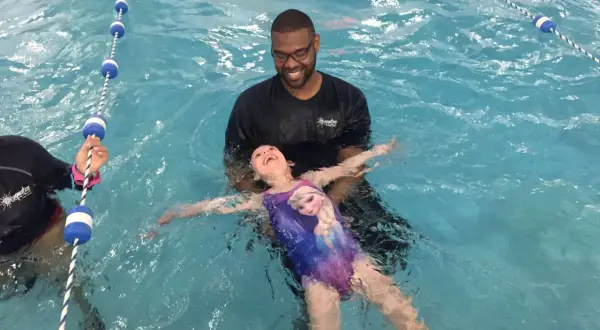 A smiling instructor helps a young girl in a Frozen swimsuit learn how to float on her back in a swimming pool.