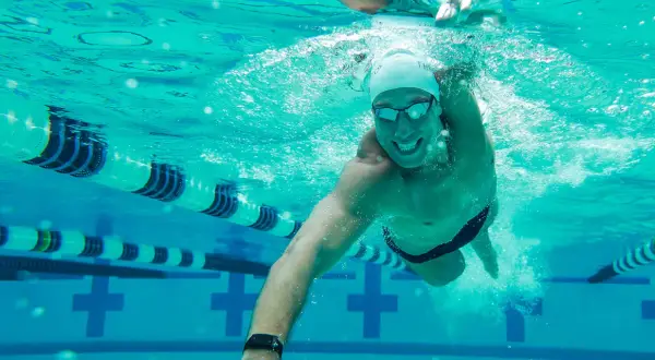 A smiling man swims underwater in a pool, enjoying easy swim workouts. Lane lines and pool markings are visible.