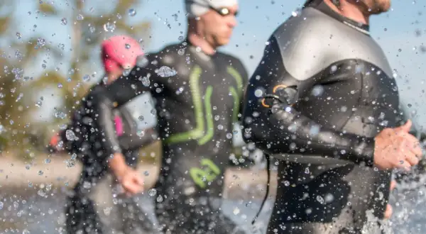 Triathletes in Ironman wetsuits run through water, splashes obscuring the view. Following Ironman wetsuit rules, they prepare for the race.