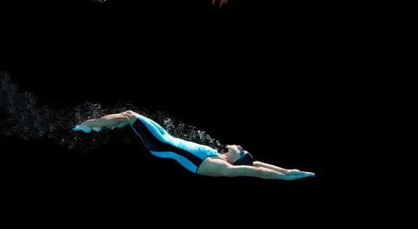 Swimmer in a blue and black suit performs a powerful dolphin kick underwater, creating a trail of bubbles against a dark background.