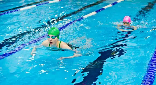 Two swimmers in a pool, one in a green cap doing breaststroke, the other in a pink cap swimming freestyle.